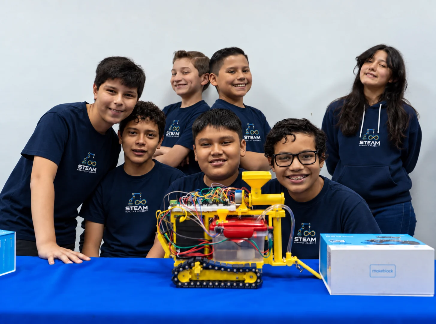 Robotics Club students smiling behind the robot they built, wearing the navy academy tee and hoodie